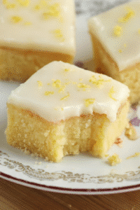 Square lemon bars with white glaze and lemon zest, one piece with a bite taken.