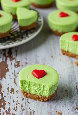 Grinch Mini Cheesecakes with green filling, whipped cream, and red heart candies in muffin liners.